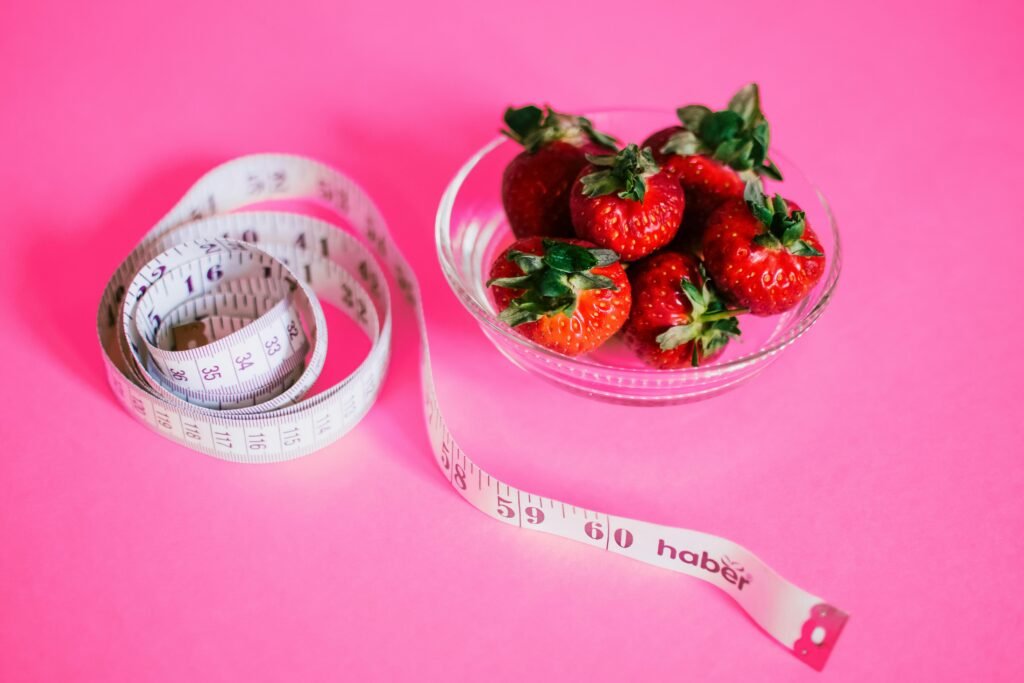 A glass bowl of fresh strawberries paired with a pink measuring tape symbolizes healthy eating.. weight management