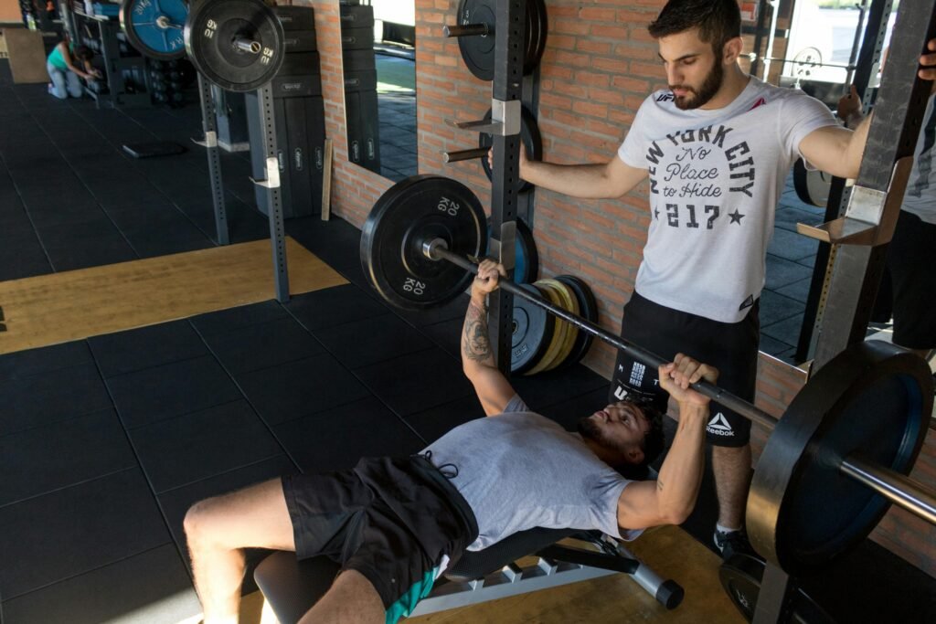 A strong man lifts weights on a bench while a trainer assists in a modern gym setup.
bench press
