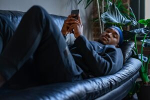 A man lying on a sofa in a comfortable indoor setting, using a smartphone.