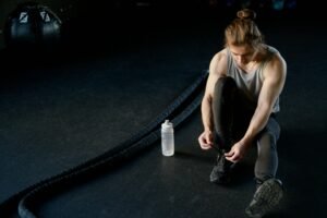 Man tying shoes on gym floor with battle ropes and water bottle, ready for training.