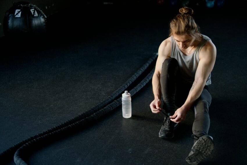 Man tying shoes on gym floor with battle ropes and water bottle, ready for training.