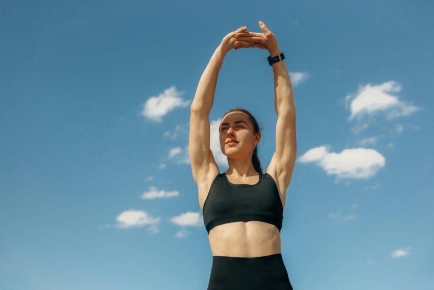 Woman in sports bra stretching under a clear blue sky, embodying fitness and wellness.